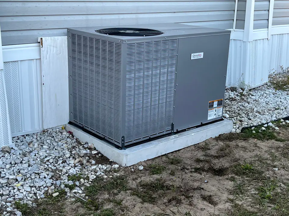 Gray package AC unit on a new concrete pad beside a manufactured home